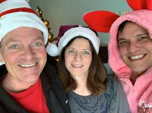 Husband and wife in their 50's with son in his 30's, in Santa Claus caps, smiling at the camera.